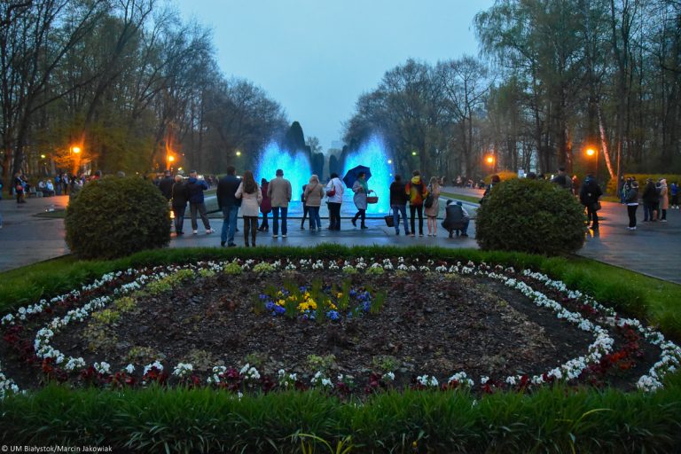 Park Plany w Białymstoku. Na pierwszym planie jest kompozycja z posadzonych kwiatów. W oddali widoczne są kolorowe fontanny, przy których stoją ludzie