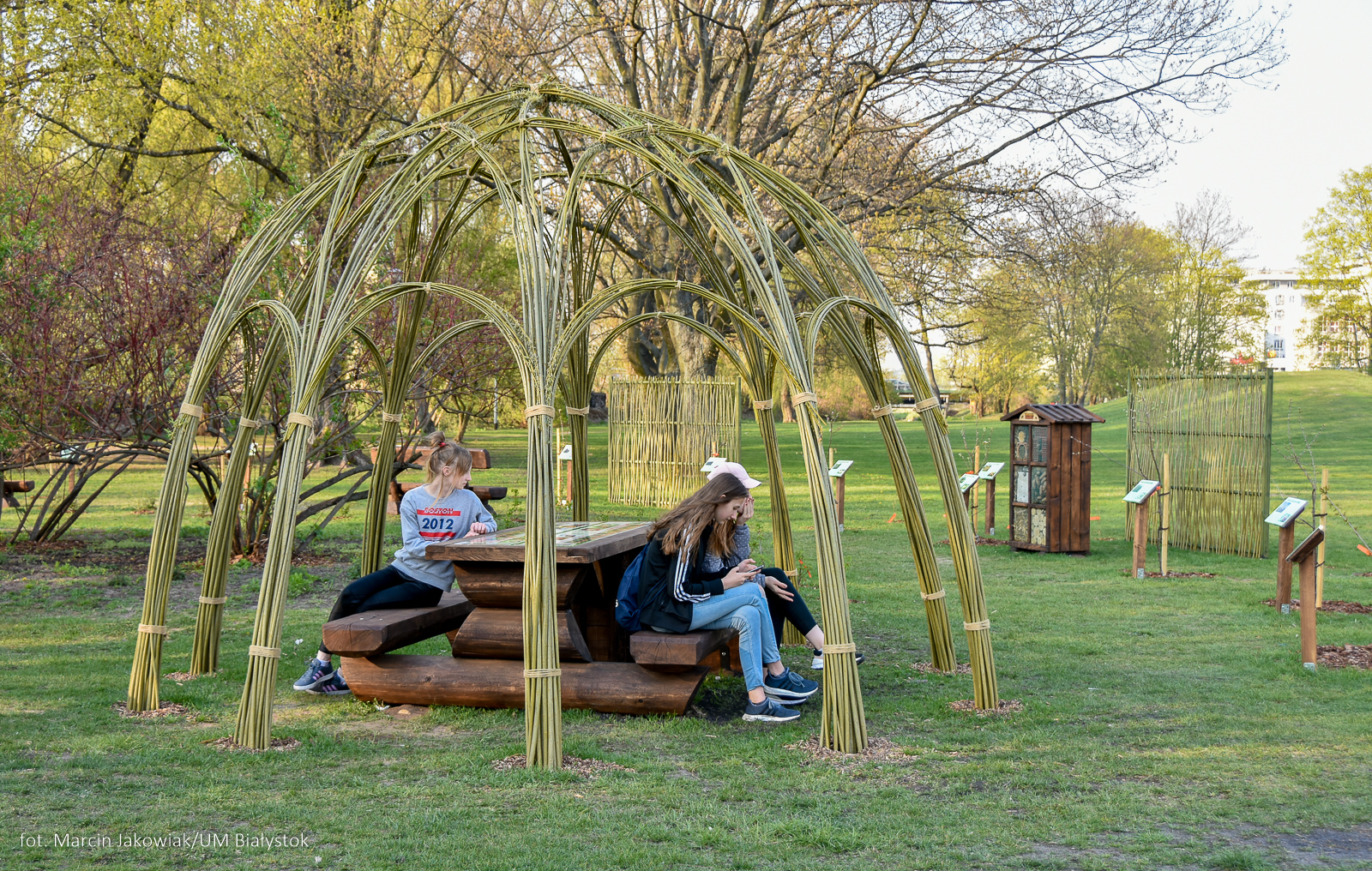 Park, ludzie siedzą na ławkach, pod konstrukcją zrobioną z traw, gałęzi