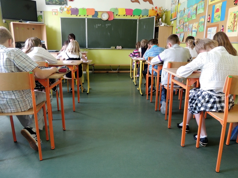 Klasa, w ławkach siedzą uczniowie, nauczycielka pisze przy tablicy
