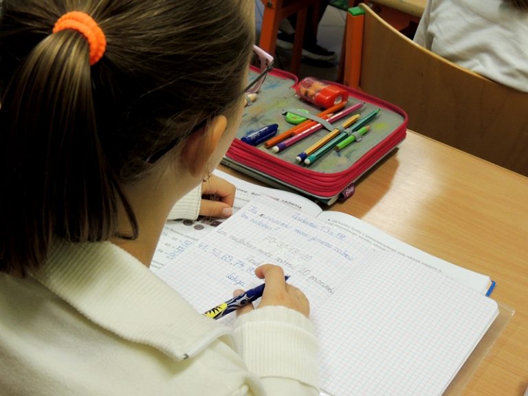 Uczennica siedzi w ławce i pisze w zeszycie. Na ławce, oprócz zeszytu, leży też piórnik