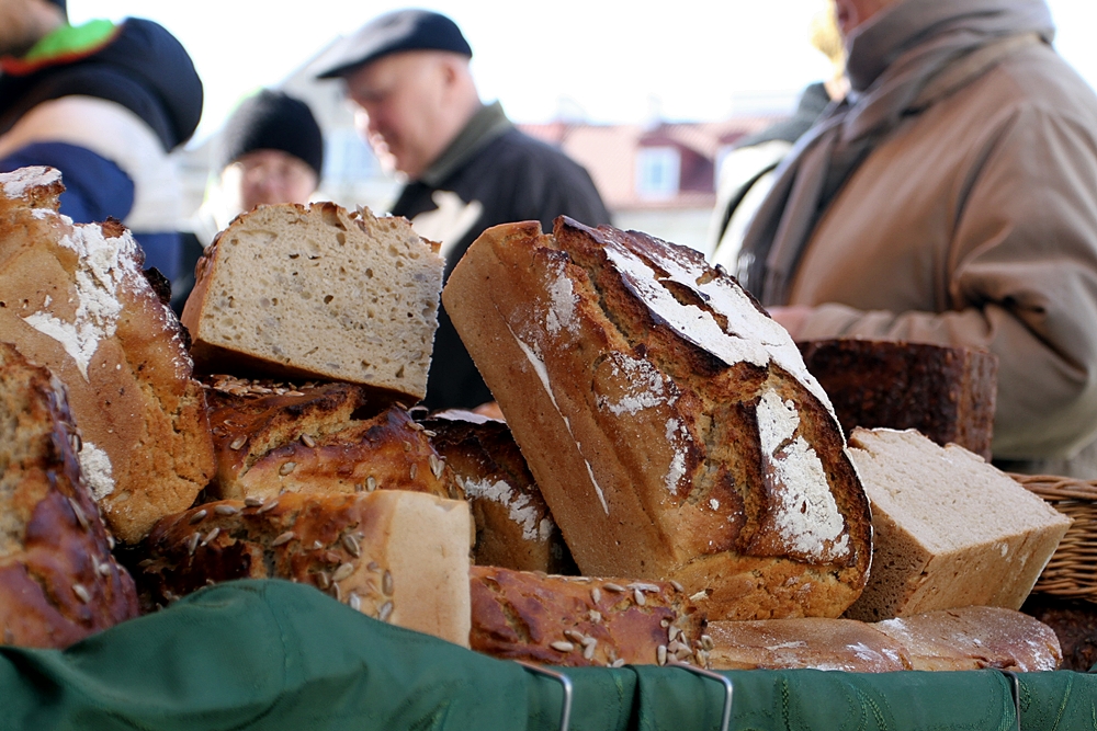 Na pierwszym planie w wiklinowym koszu leżą bochenki chleba, w tle widać kupujących ludzi. Rzecz dzieje się na podwórku, przy stoiskach na Jarmarku