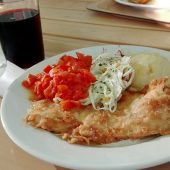 Na białym talerzu leży kotlet schabowy w panierce, ziemniaki, surówki. Obok talerza stoi szklanka z napojem