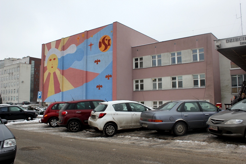 Część budynków Uniwersyteckiego Dziecięcego Szpitala Klinicznego w Białymstoku. Na budynku, z lewej strony, namalowane jest słońce. Przed szpitalem stoją zaparkowane samochody