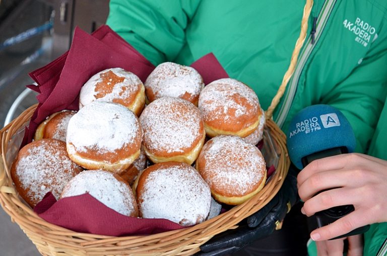 Pączki w pudrze leżą w wiklinowym koszyku, na bordowej serwetce. Widać fragment zielonej kurtki osoby, która trzyma koszyk z pączkami