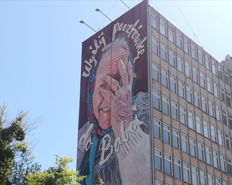Wysoki budynek, na którego bocznej ścianie znajduje się mural. Malowidło przedstawia straszą panię w chustce na głowie, która dłonią zasłania twarz. Na muralu znajduje się napis: Wyślij pocztówkę do babci.