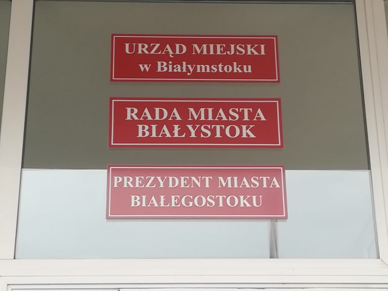 Trzy czerwone tabliczki na drzwiach budynku magistratu. Na pierwszej napisano Urząd miejski w Białymstoku, na drugiej - Rada Miasta Białystok, a na trzeciej - Prezydent Miasta Białegostoku