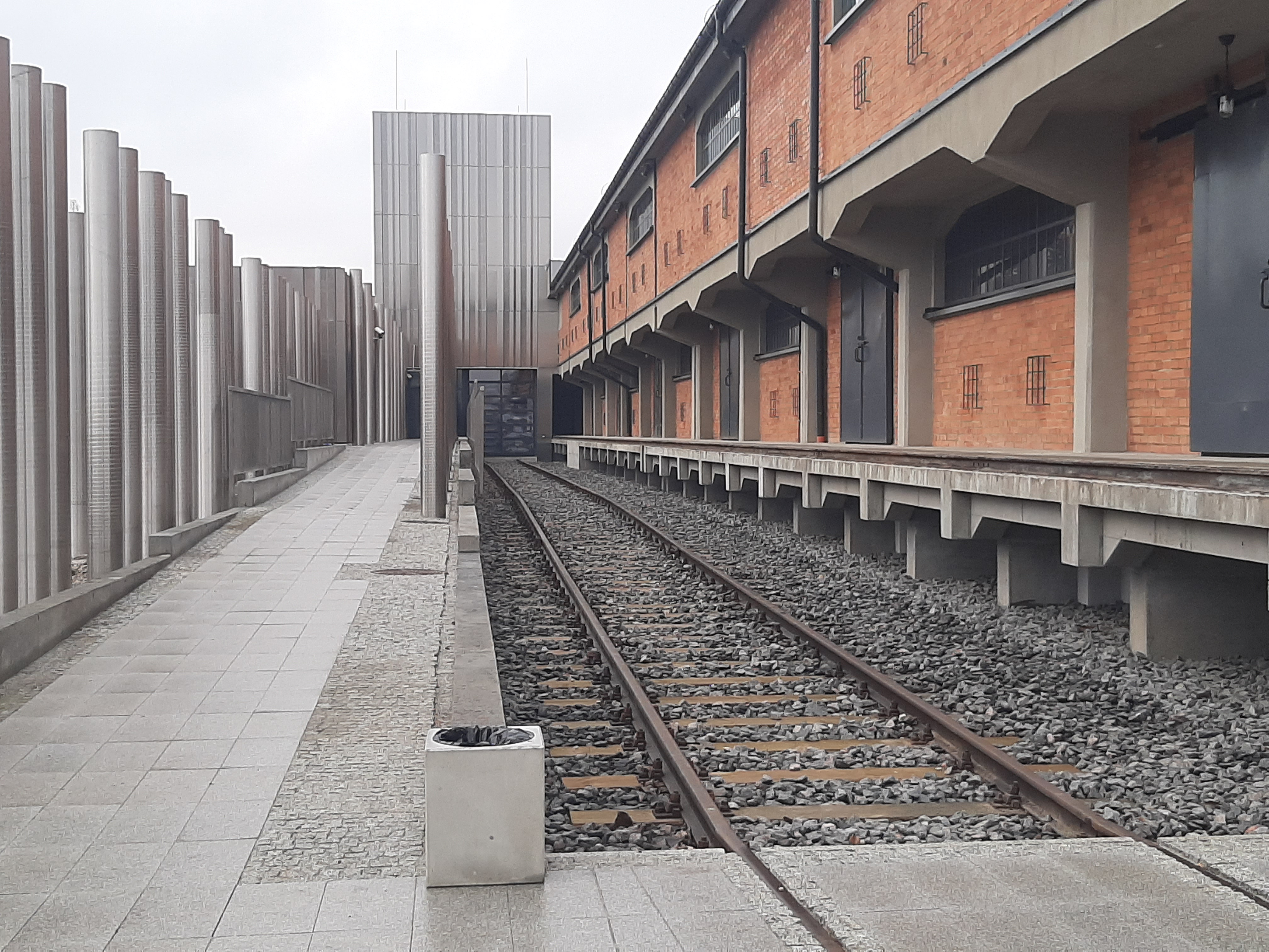 Droga prowadząca do wejścia głównego Muzeum Pamięci Sybiru. Po prawej stronie widoczne są ceglane budynki oraz tory kolejowe. Po lewej stronie są aluminiowe tuby symbolizujące tajgę