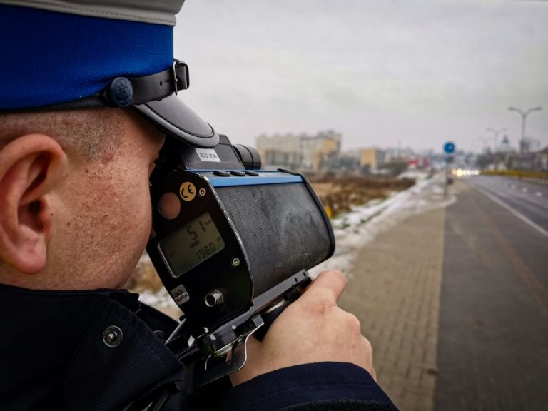 Policjant trzyma przy twarzy prędkościomierz, po prawej stronie widać ulicę