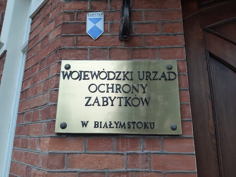 Do ceglastej ściany przyczepiona jest miedziana tabliczka, na której napisane jest Wojewódzki Urząd Ochrony Zabytków