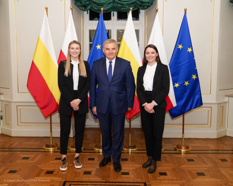 Po środku stoi prezydent Białegostoku, po jego prawej stronie znajduje się Natalia Maliszewska, a po lewej Patrycja Maliszewska. Dziewczyny są ubrane w czarne spodnie, białe koszule i czarne marynarki. Prezydent jest w granatowym garniturze. Za postaciami są ustawione flagi Białegostoku, Polski i Unii Europejskiej