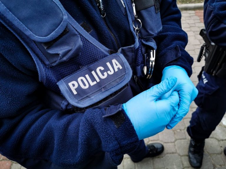 Policjant stoi w pełnym umundurowaniu. Ma na rękach jednorazowe rękawiczki ochronne.