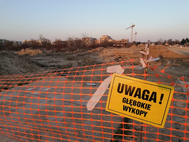 na ogrodzeniu z siatki wisi znak Uwaga głębokie wykopy, za siatką miejsce pod budowę