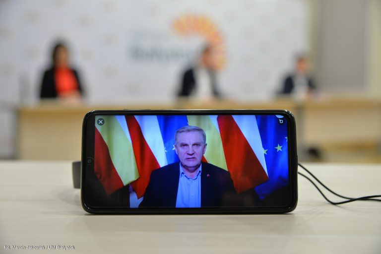Telefon komórkowy stoi na biurku, na wyświetlaczu widać twarz prezydenta Białegostoku