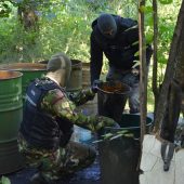 Funkcjonariusze KAS stoją na terenie leśnej bimbrowni. Na miejscu jest kadź zawierająca 300 litrów fermentującego zacieru, chłodnica i kocioł, a także doły z wodą wykorzystywaną w wytwarzaniu bimbru oraz rozlewisko