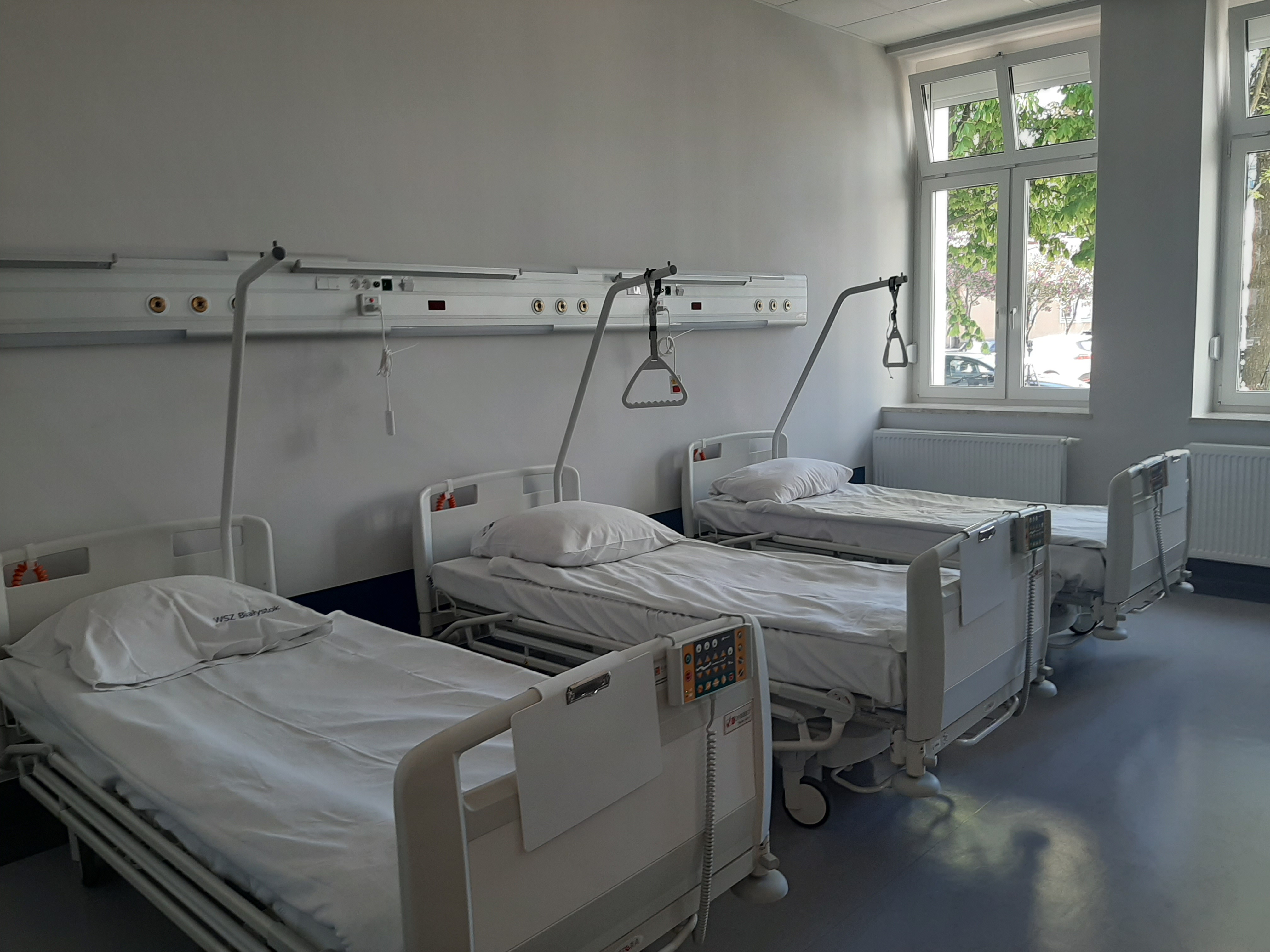 W sali szpitalnej stoją trzy łóżka, na których znajduje się biała pościel. Po prawej stronie jest okno