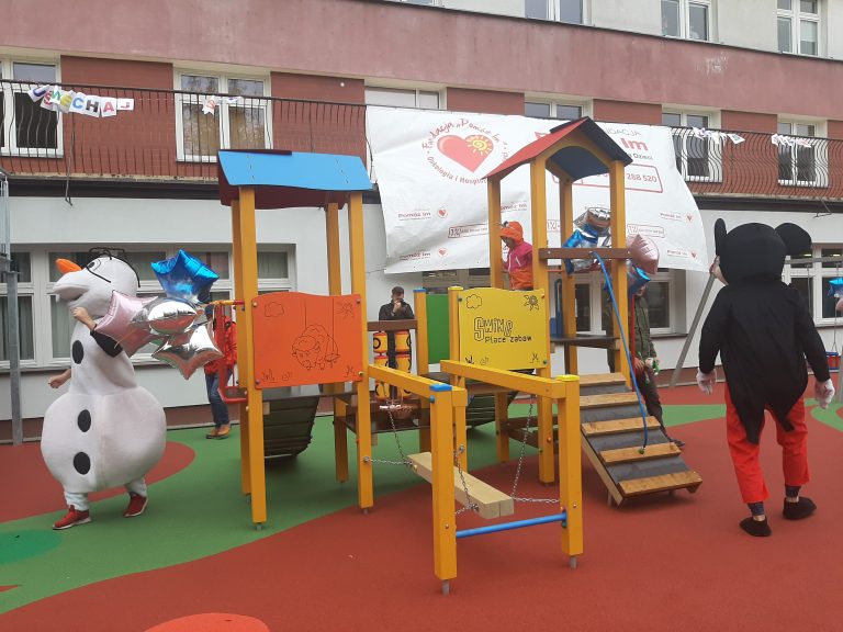 Plac zabaw przed oddziałem onkologii UDSK. Dzieci biegają po drabinkach. Z lewej strony jest chodząca maskotka bałwan, z prawej zaś tyłem odwrócona Myszka Miki