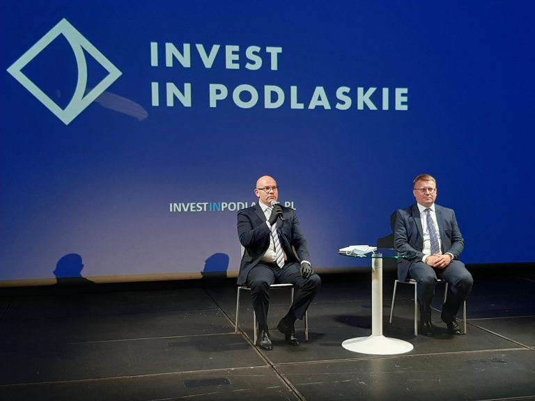 Dwóch mężczyzn, w tym marszałek siedzą przy okrągłym, białym stoliku. Za ich plecami na granatowym tle jest biały napis Invest In Podlaskie. Marszałek, który znajduje się po lewej stronie zdjęcia mówi przez mikrofon