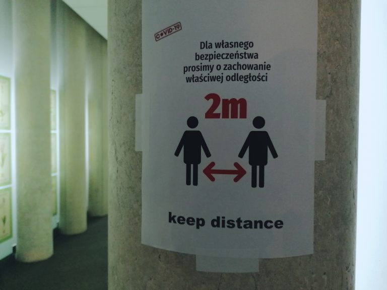 Na słupie w korytarzu wisi plakat przypominający o obowiązku zachowania dystansu społecznego.