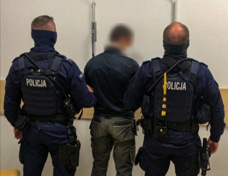 2 policjantów w mundurach, którzy trzymają zatrzymanego mężczyznę.