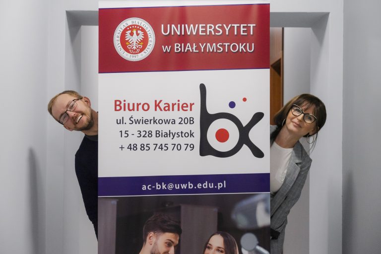 Mężczyzna i kobieta wyglądają zza roll-upu reklamującego Biuro Karier Uniwersytetu w Białymstoku.