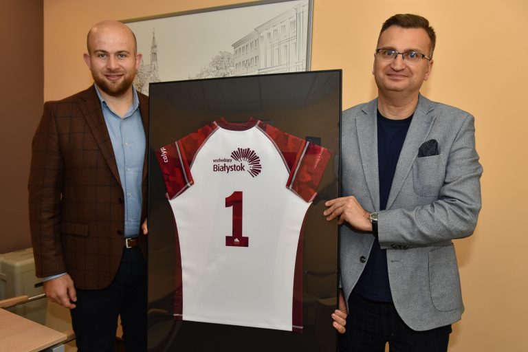 Po lewej stronie stoi mężczyzna ubrany w granatowe spodnie, jasno niebieską koszulę i bordową marynarkę. Po prawej stronie stoi zastępca prezydenta Białegostoku ubrany w ciemną koszulkę i szarą marynarkę. Panowie trzymają białą z czerwonymi rękawami koszulkę do gry w rugby. T-shirt to prezent od drużyny rugby dla wiceprezydenta