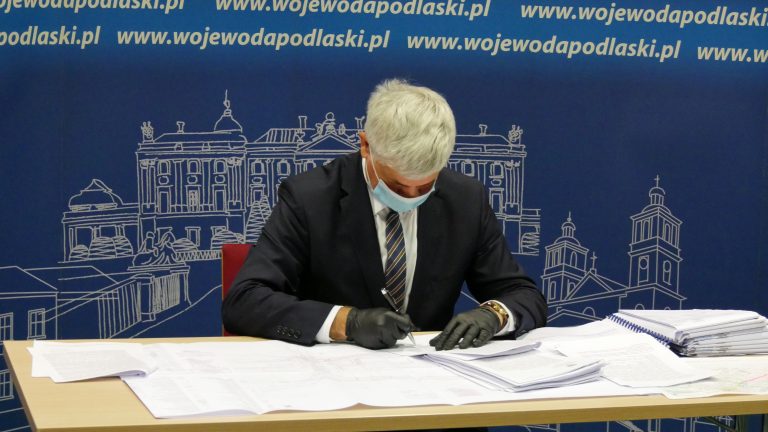 Wojewoda pochylony nad planami rozbudowy tras kolejowych podpisuje pozwolenie na kolejną rozbudowę odcinka. Wojewoda jest ubrany w czarny garnitur, za nim niebieska ścianka promocyjna Podlaskiego Urzędu Wojewódzkiego w Białymstoku