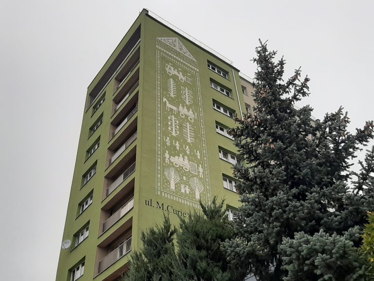 Na tle szarego nieba znajduje się zielony wieżowiec. Na jego ścianie widoczny jest biały mural przedstawiający fragment tkaniny dwuosnowowej. Budynek nieco przysłania drzewo iglaste