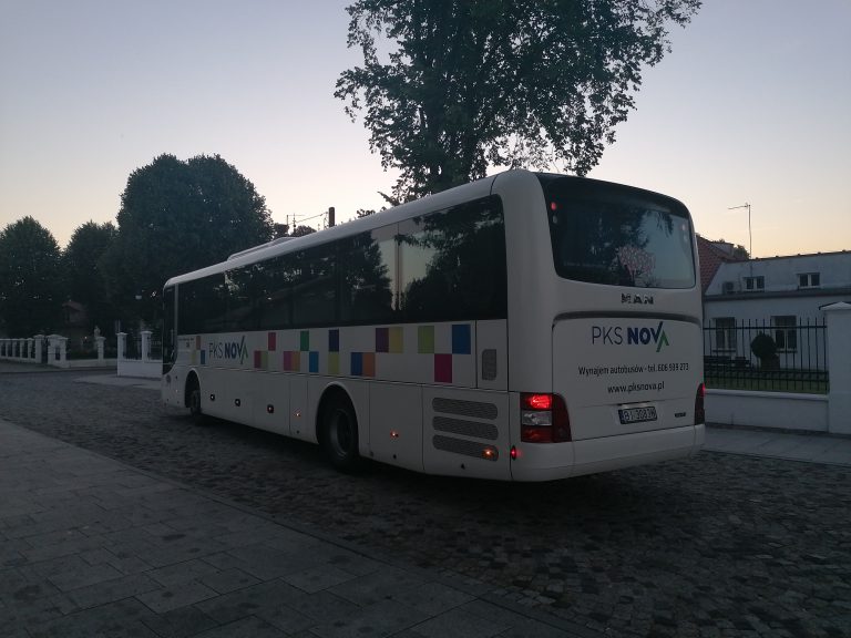 Na brukowanej ulicy stoi biały, w kolorowe kwadraty autobus, typu autokar. Pojazd stoi tyłem, widoczna jest tylna szyba i jego boczna ściana