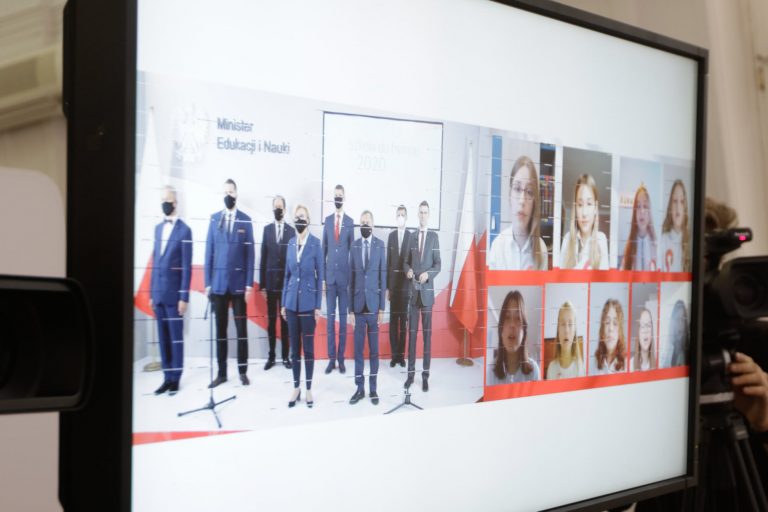 Na ekranie telewizora po lewej stronie są przedstawiciele Ministerstwa Edukacji i Nauki. Po prawej stronie ekranu widocznych jest 9 dziewczynek, która każda ze swojego domu za pośrednictwem komputera będzie śpiewała hymn