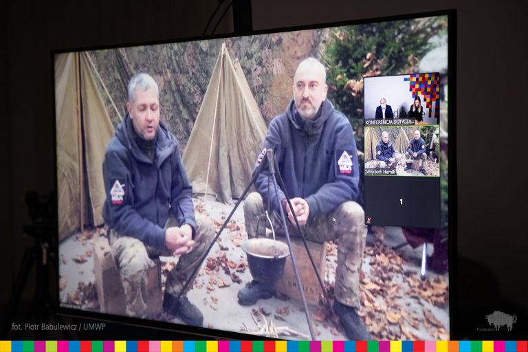 Na monitorze widocznych jest dwóch mężczyzn, którzy siedzą w zrekonstruowanym obozowisku harcerskim. Mężczyźni siedzą wokół paleniska, za nimi widoczne są namioty