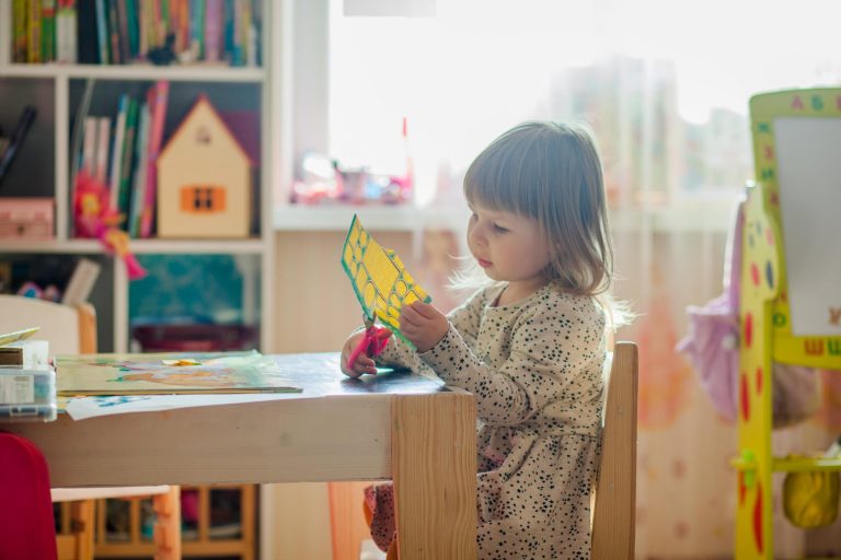 Dziecko siedzi na krzesełku przy stoliku i wycina nożyczkami z kartki papieru. Dziewczynka znajduje się pokoju wypełnionym zabawkami