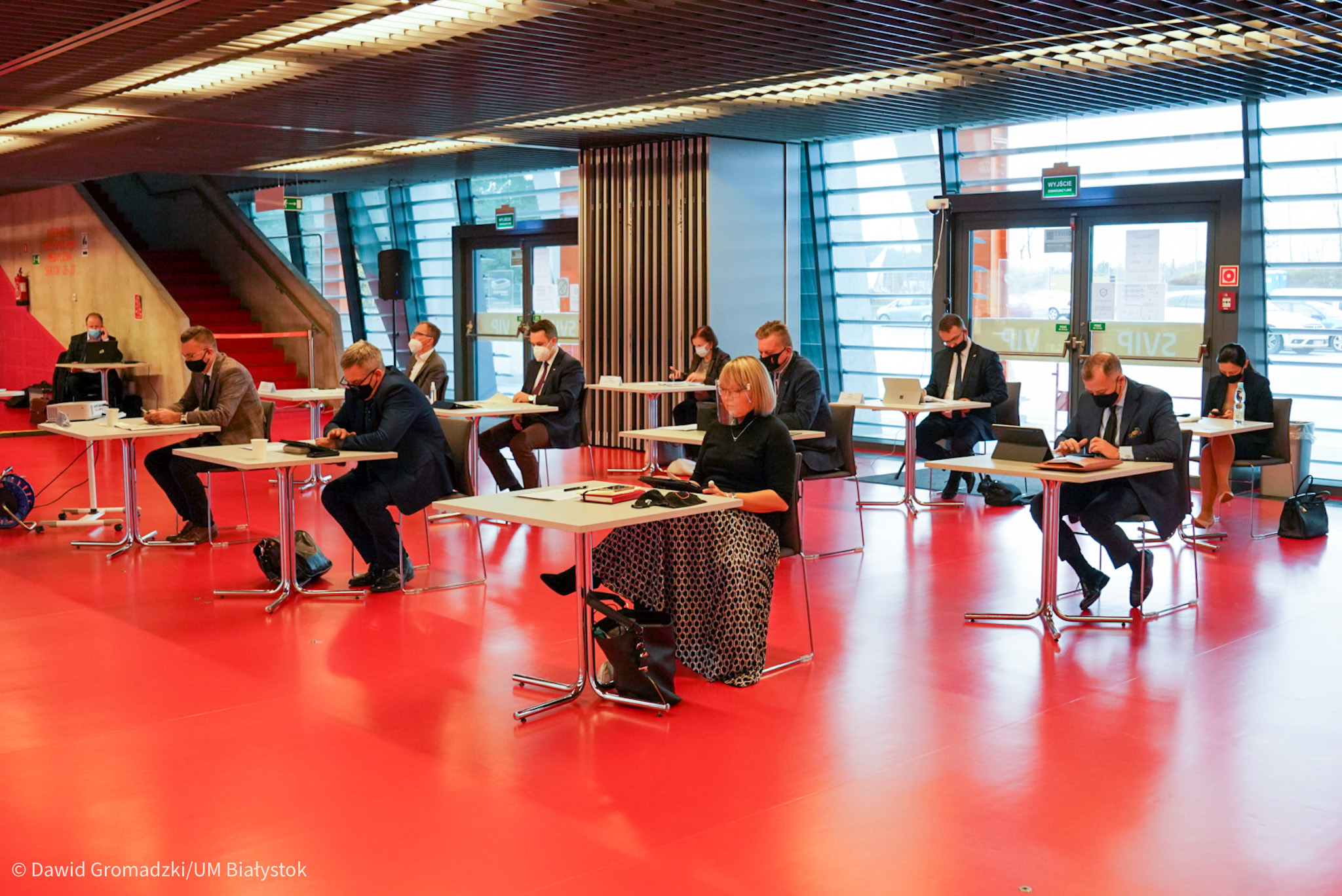 W sali konferencyjnej Stadionu Miejskiego w Białymstoku, gdzie jest intensywnie czerwona podłoga odbywa się sesja miasta. Radni siedzą pojedynczo przy stolikach, które stoją od siebie w odległości około 2 metrów