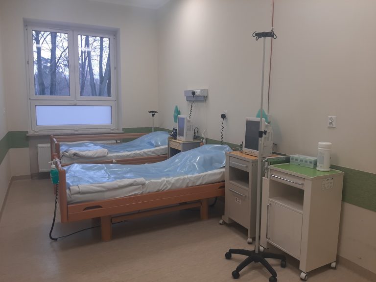 Sala szpitala, stoją dwa łóżka, dwie szafki, stojak na kroplówkę