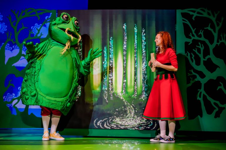 Na scenie teatralnej stoi aktor przebrany w dużą, zieloną żabę, obok stoi rudowłosa aktorka ubrana w czerwoną sukienkę