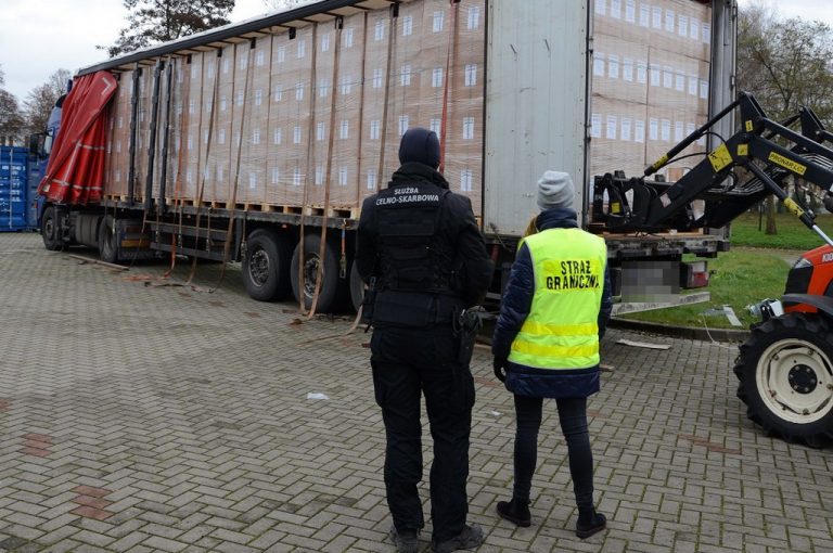 Ciężarówka wypchana nielegalnymi papierosami. Tirowi przygląda się dwóch funkcjonariuszy, stojący po lewej stronie jest ubrany na czarno, a osoba po prawej stronie ma na sobie zieloną odblaskową kamizelkę