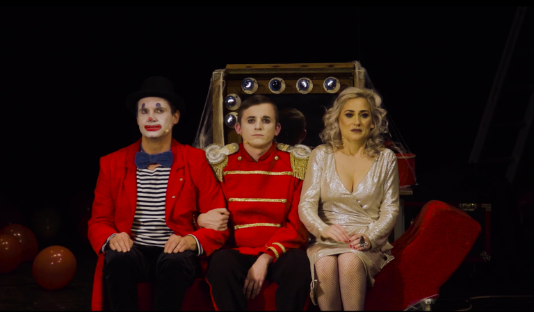 Na czerwonej kanapie, od lewej strony siedzi clown, pośrodku mężczyzna w czerwonej marynarce, po prawej kobieta w złotej sukience
