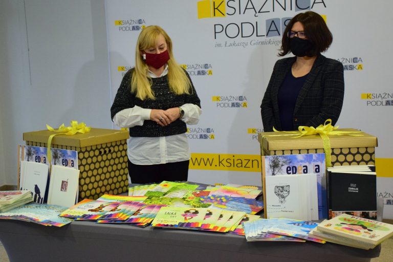 Dwie kobiety stoją za stołem, na którym ułożone są książki. Kobieta po lewej stronie ma długie, blond włosy i jest ubrane w czarne spodnie, białą koszulę i czarny sweterek. Kobieta po prawej stronie ma krótkie ciemne włosy, jest również ubrana na czarno. Za kobietami stoi baner z napisem Książnica Podlaska