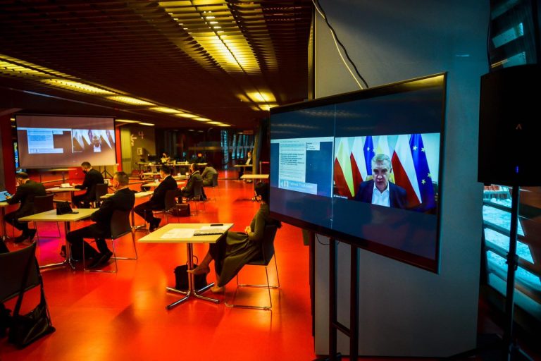 Sala konferencyjna Stadionu Miejskiego w Białymstoku. Po lewej stronie, przy pojedynczych stolikach siedzą radni, po prawej stronie jest ustawiony duży monitor, na którym widoczny jest prezydent Białegostoku