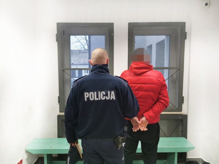 Pomieszczenie budynku komendy, policjant z mężczyzną, odwróceni tyłem, patrzący w kierunku okna, które jest okratowane