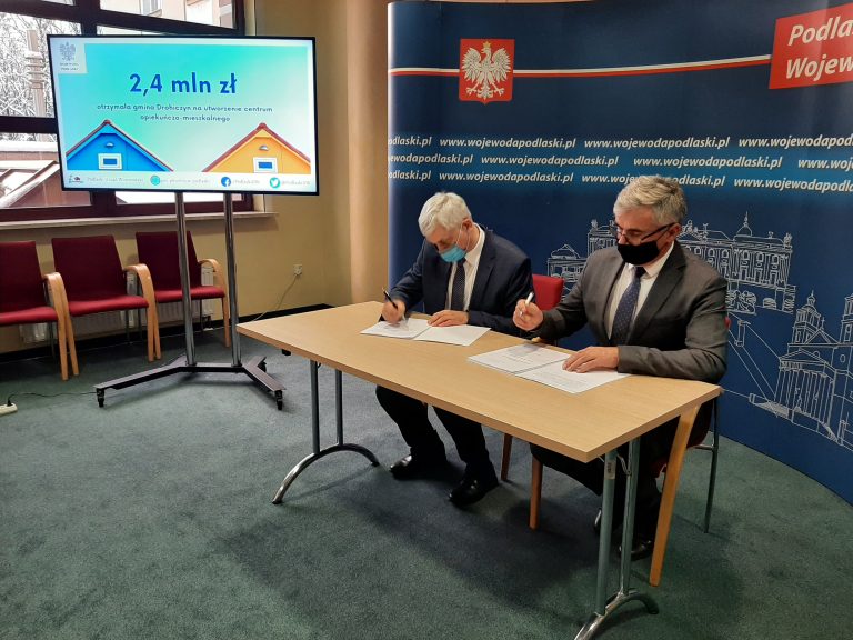 Wojewoda i burmistrz gminy Drohiczyn podpisują umowę. Mężczyźni siedzą przy stole, za nimi znajduje się ścianka Podlaskiego Urzędu Wojewódzkiego w Białymstoku. W lewym rogu zdjęcia widoczny jest duży telewizor, na którym jest wyświetlona kwota 2.4 miliona złotych.