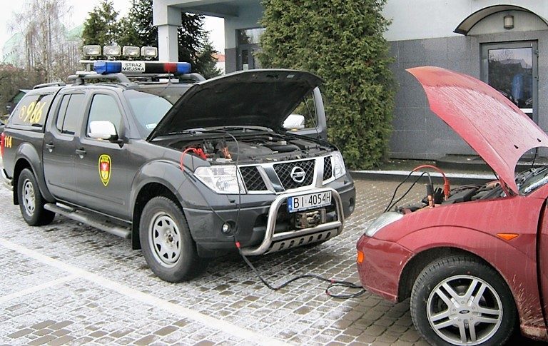 Dwa samochody stoją zwrócone do siebie przodem. Są podłączone kablami rozruchowymi