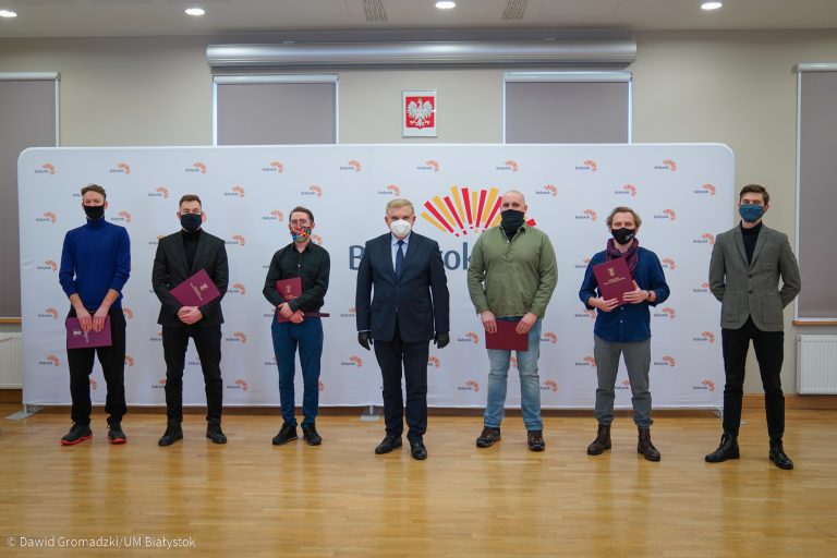 Na tle ścianki Wschodzącego Białegostoku stoi siedem osób. Sami mężczyźni. Po środku stoi prezydent Białegostoku, a po jego obu stronach stypendyści, którzy otrzymali dotację na działania artystyczne
