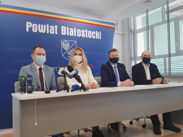 Cztery osoby, trzech mężczyzn i jedna kobieta siedzą przy stole. Za nimi znajduje się ścianka z napisem Powiat Białostocki