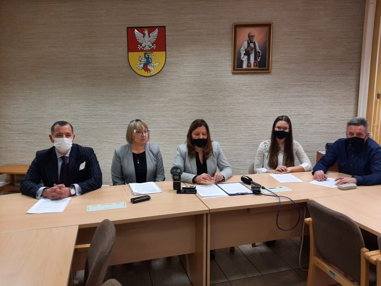 Pięciu radnych siedzi przy stole, po środku kobiety, bo bokach mężczyźni. Za nimi na ścianie wisi herb Białegostoku oraz wizerunek patrona Białegostoku błogosławionego księdza Michała Sopoćki