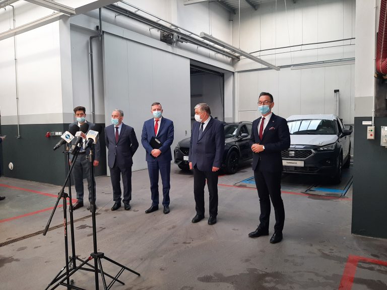 Pięciu mężczyzn stoi w hali, w której serwisowane i naprawiane są samochody. Za nimi stoją dwa uta, natomiast przed nimi mikrofony, po trwa konferencja prasowa
