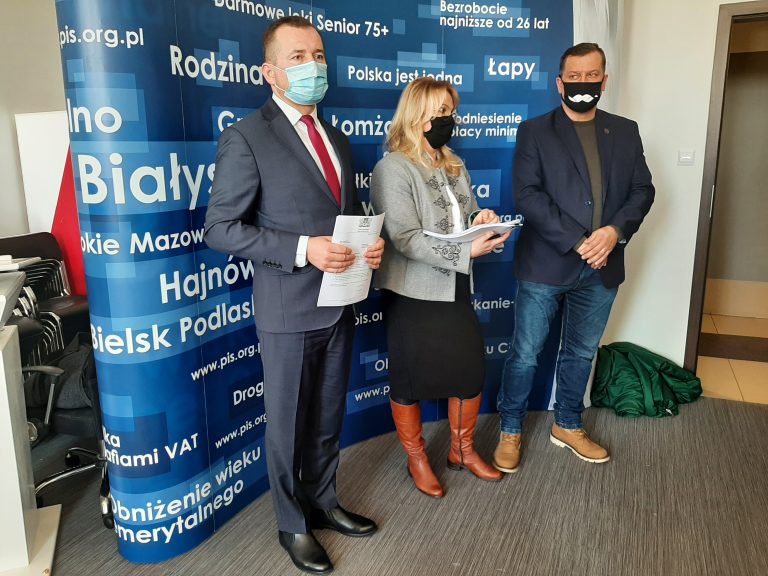 Na niebieskiej ściance w różne białe napisy stoją trzy osoby, po środku jest kobieta