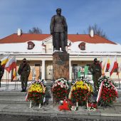 Jest słoneczny dzień, w centralnym punkcie zdjęcia znajduje się pomnik Józefa Piłsudskiego, po bokach stoją żołnierze. Za pomnikiem stoi kremowy budynek, dawna siedziba archiwum państwowego. Przed pomnikiem leżą wieńce