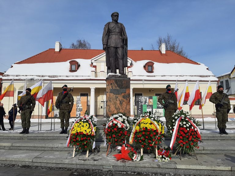 Jest słoneczny dzień, w centralnym punkcie zdjęcia znajduje się pomnik Józefa Piłsudskiego, po bokach stoją żołnierze. Za pomnikiem stoi kremowy budynek, dawna siedziba archiwum państwowego. Przed pomnikiem leżą wieńce