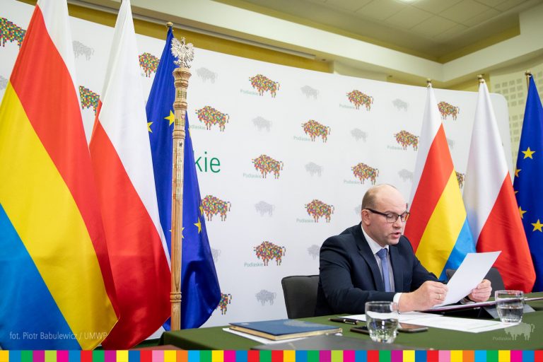 Marszałek siedzi przy stole nakrytym zielonym materiałem, za nim znajduje się ścianka, w logo - żubra, województwa podlaskiego. Po obu stronach marszałka znajdują się flagi województwa podlaskiego, Polski, Unii Europejskiej