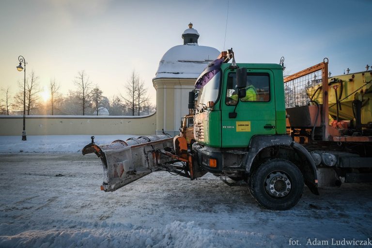 Jedzie pług zimowy, który ma zieloną kabinę. W tle widoczne jest ogrodzenie Pałacu Branickich